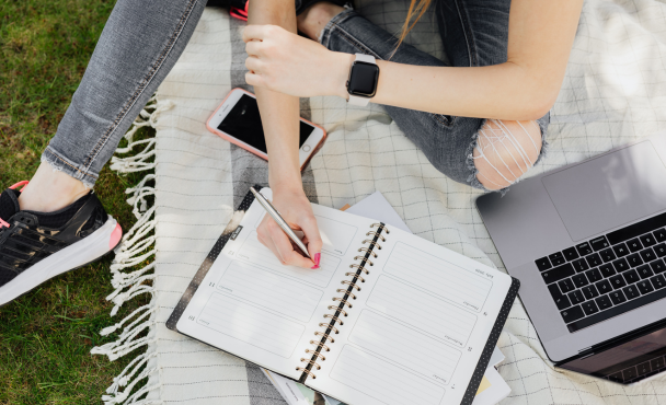 student sitting on picnic rug with laptop and mobile, writing in diary